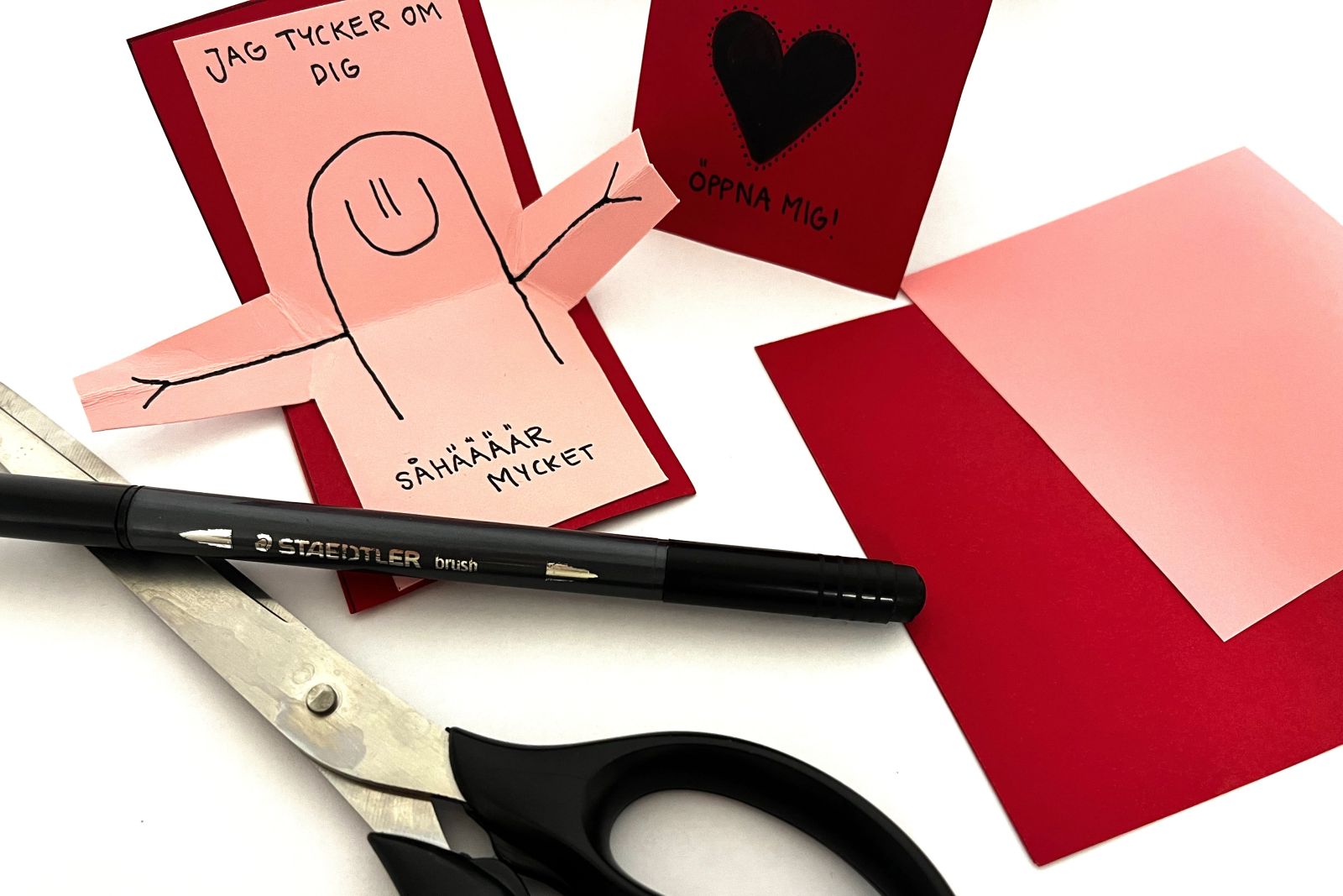 Rosa och rött alla hjärtans dag kort med texten 'jag tycker om dig såhär mycket' och en glad figur som håller ut armarna. Sax, penna och papper i bakgrunden.